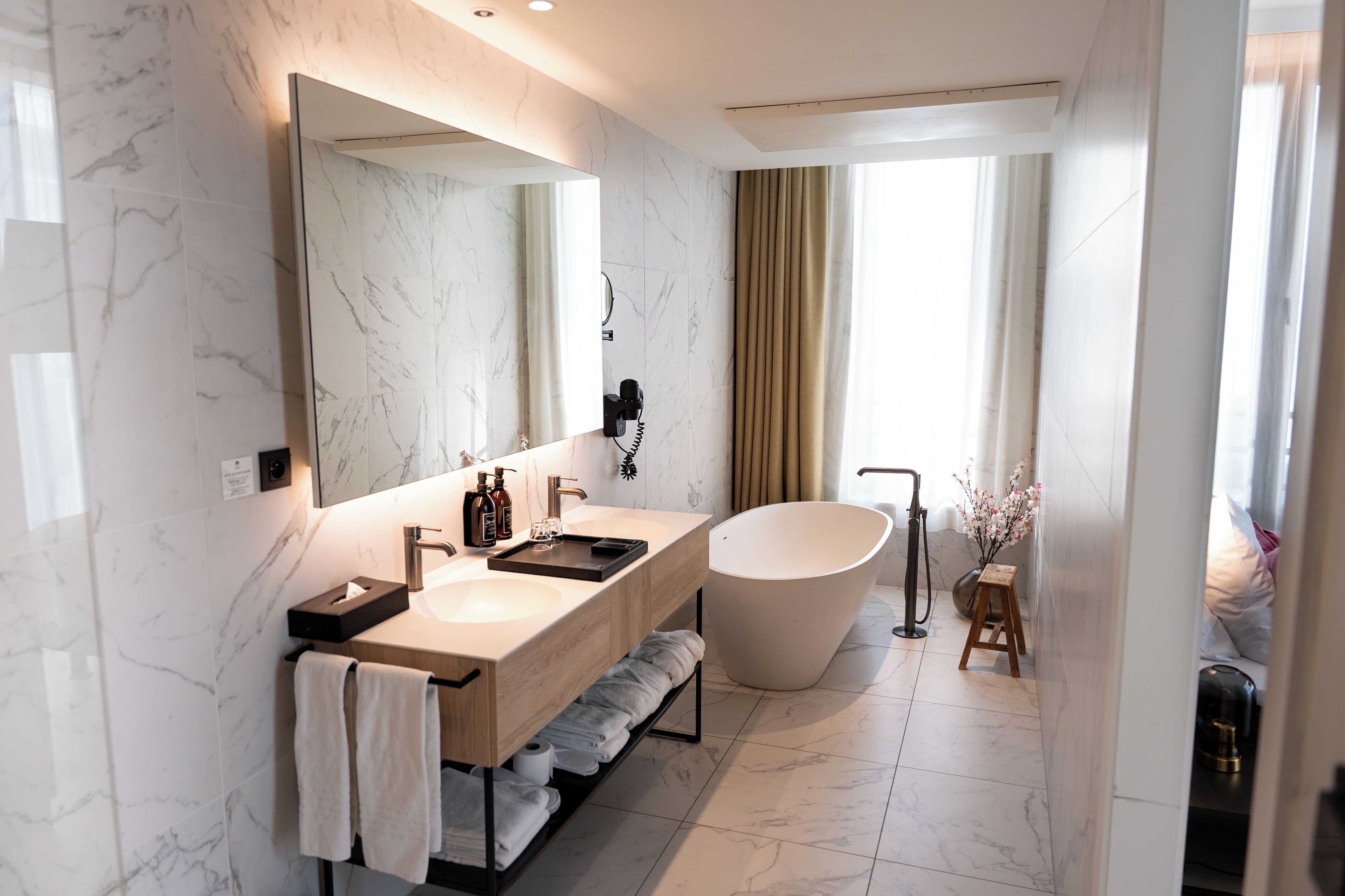 Un baño moderno con paredes de mármol, un espejo grande, una bañera independiente y un tocador con toallero.