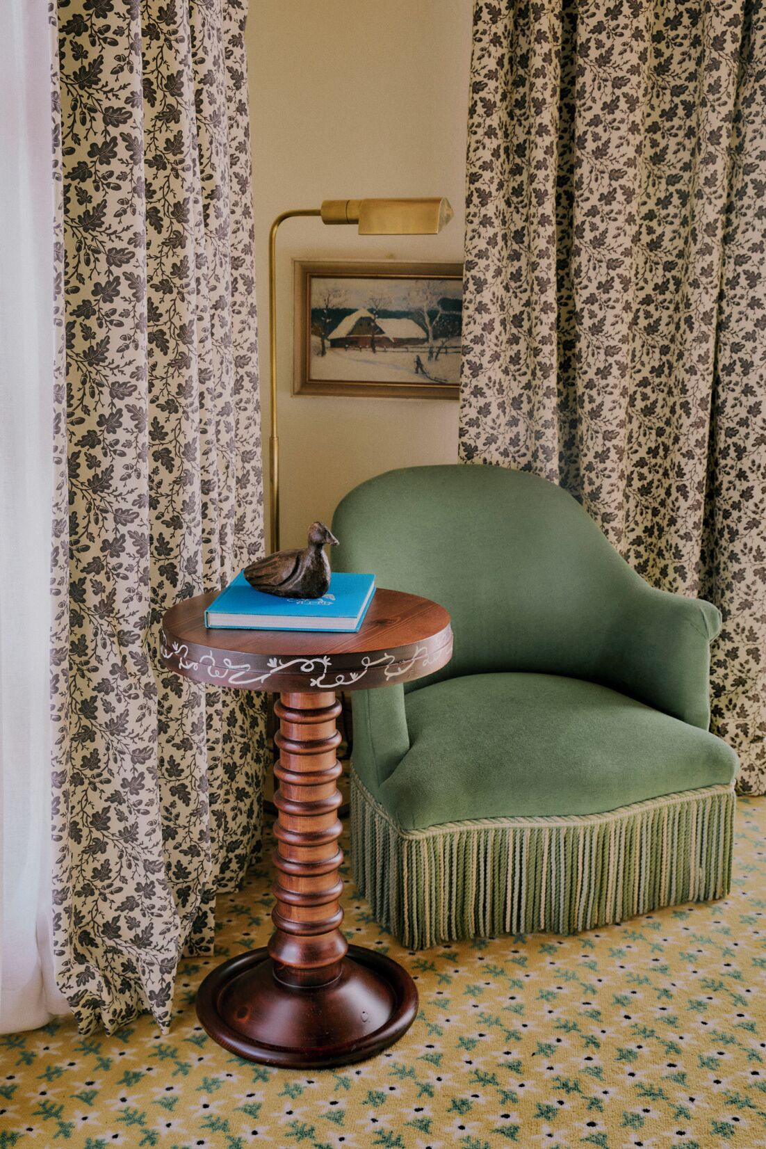A room with a green armchair, a small wooden table with a book and a figurine, floral curtains, and a framed picture on the wall.