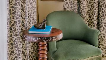 A room with a green armchair, a small wooden table with a book and a figurine, floral curtains, and a framed picture on the wall.