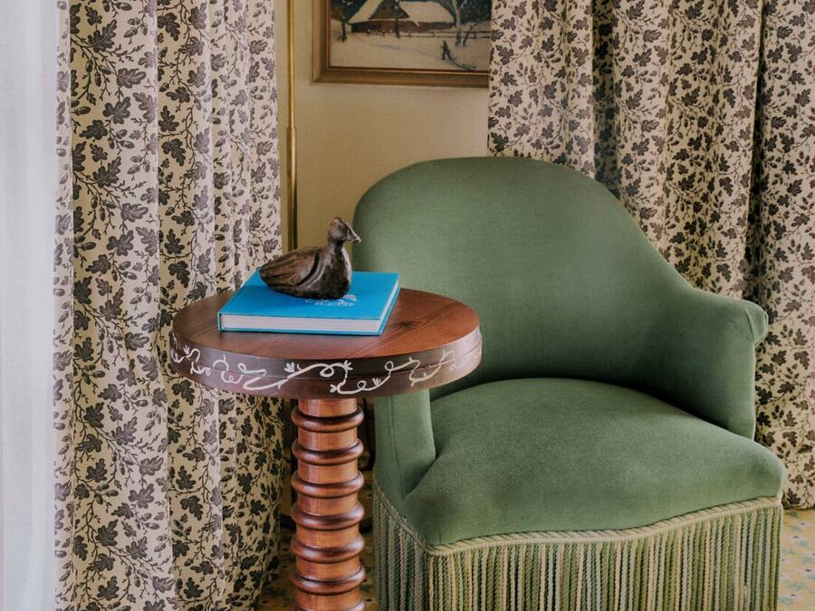 A room with a green armchair, a small wooden table with a book and a figurine, floral curtains, and a framed picture on the wall.
