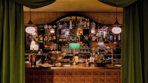 Una zona de bar con mostrador de madera, estantes con botellas de licores, una lámpara verde y un tazón grande.