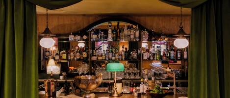 Una zona de bar con mostrador de madera, estantes con botellas de licores, una lámpara verde y un tazón grande.