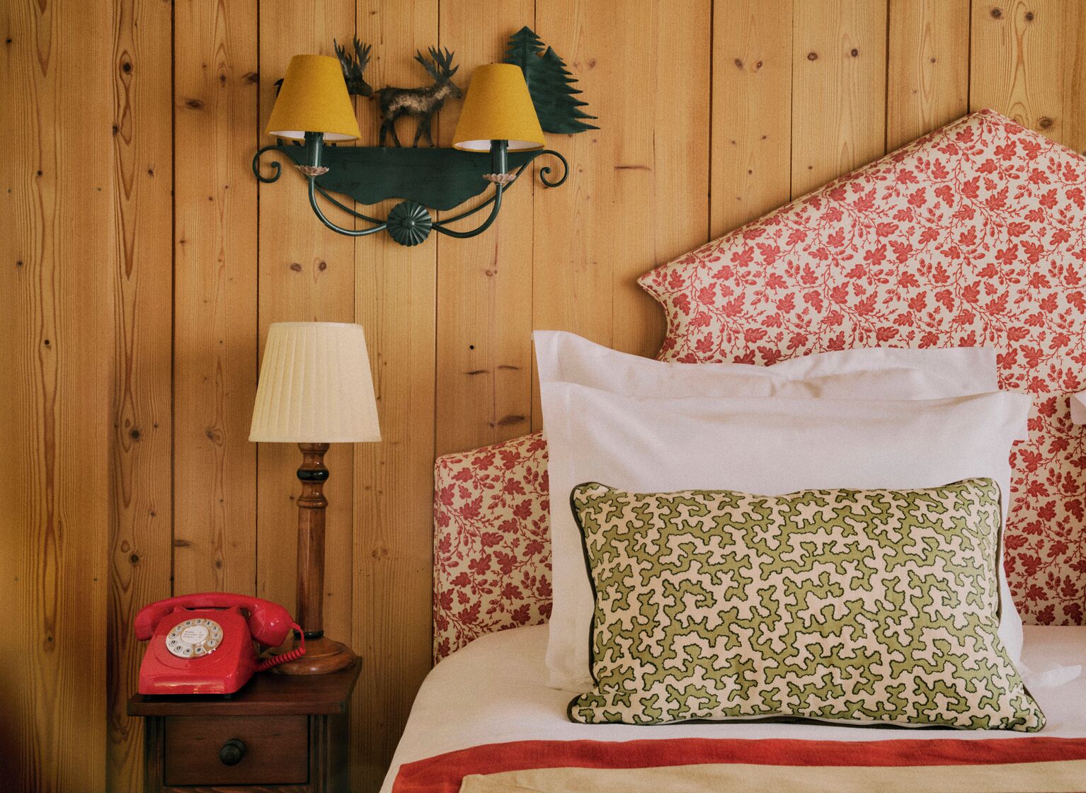 A wooden-paneled room with a bed, floral-patterned pillows, a red rotary phone, and a wall-mounted lamp with two lampshades.