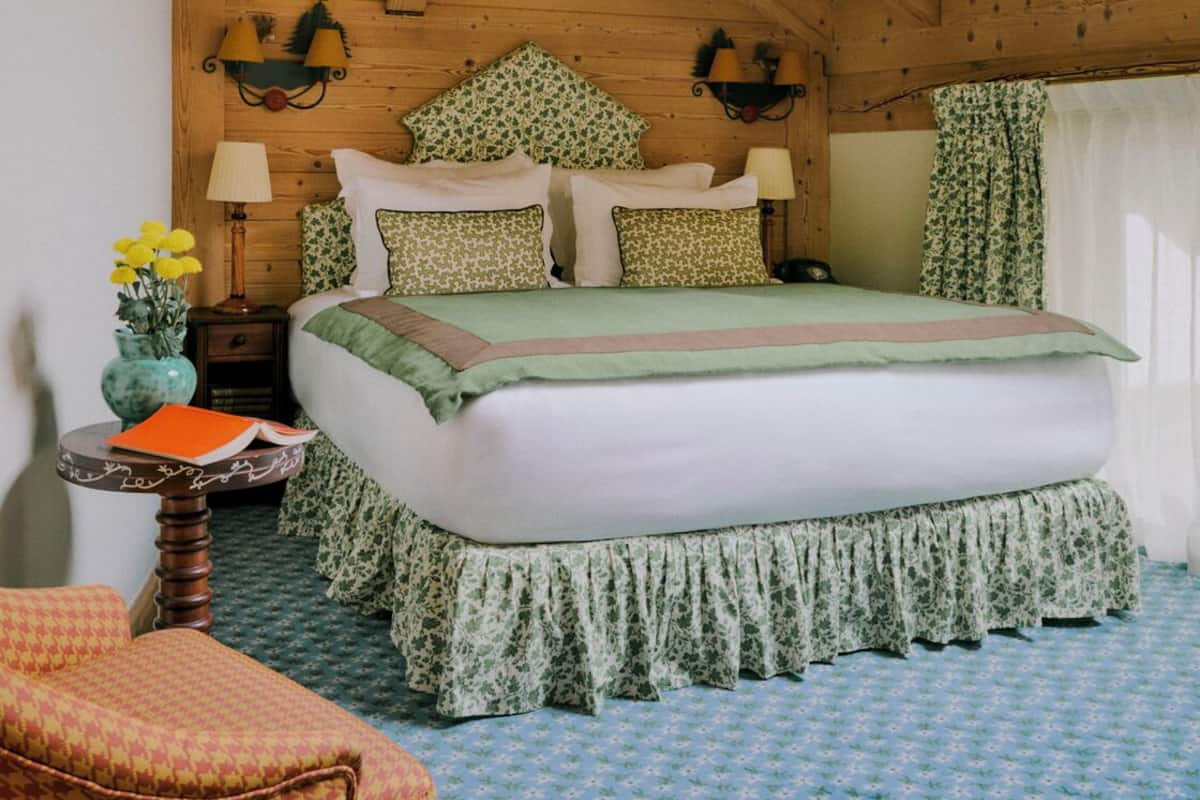 A cozy bedroom with a wooden ceiling, a bed with floral bedding, a nightstand with a lamp, and a chair.
