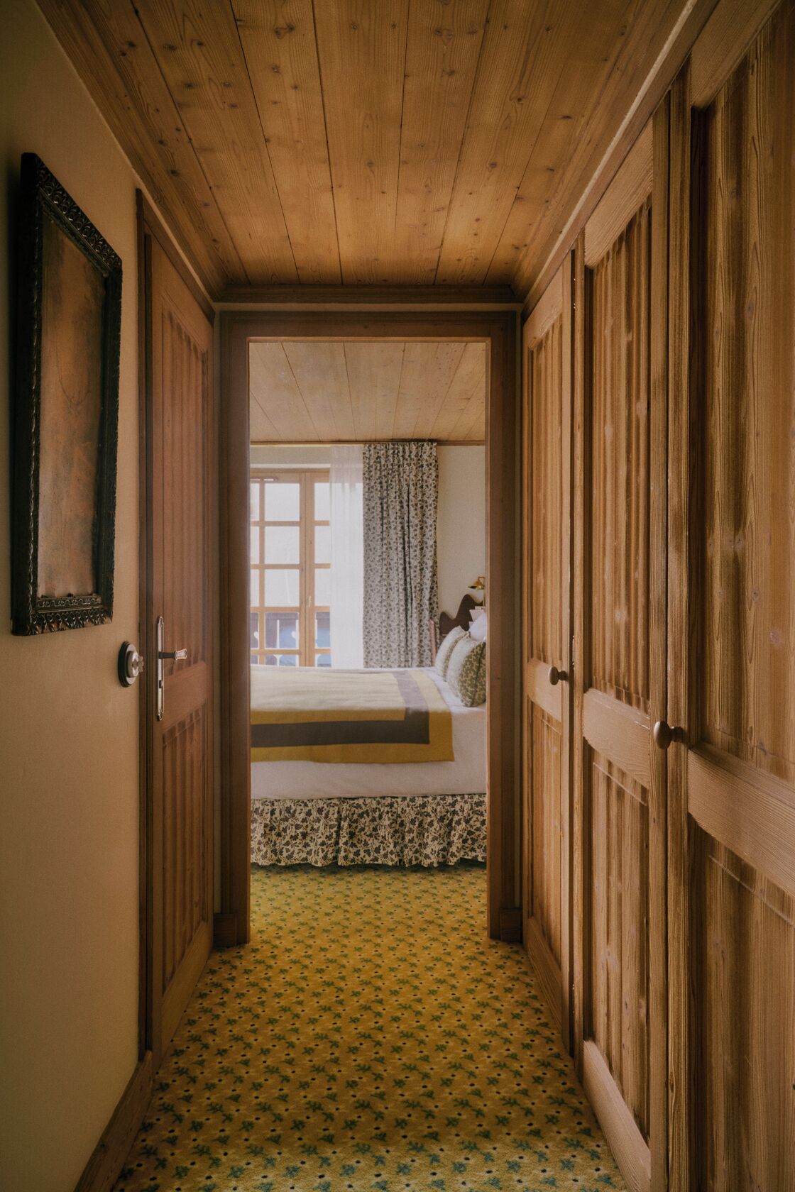 A narrow hallway with wooden doors leading to a bedroom. The room has a bed with a floral-patterned bedspread and a window with curtains.