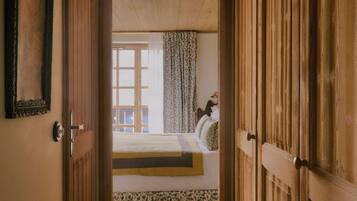 Een smalle gang met houten deuren die leiden naar een slaapkamer. De kamer heeft een bed met een bloemenprint sprei en een raam met gordijnen.