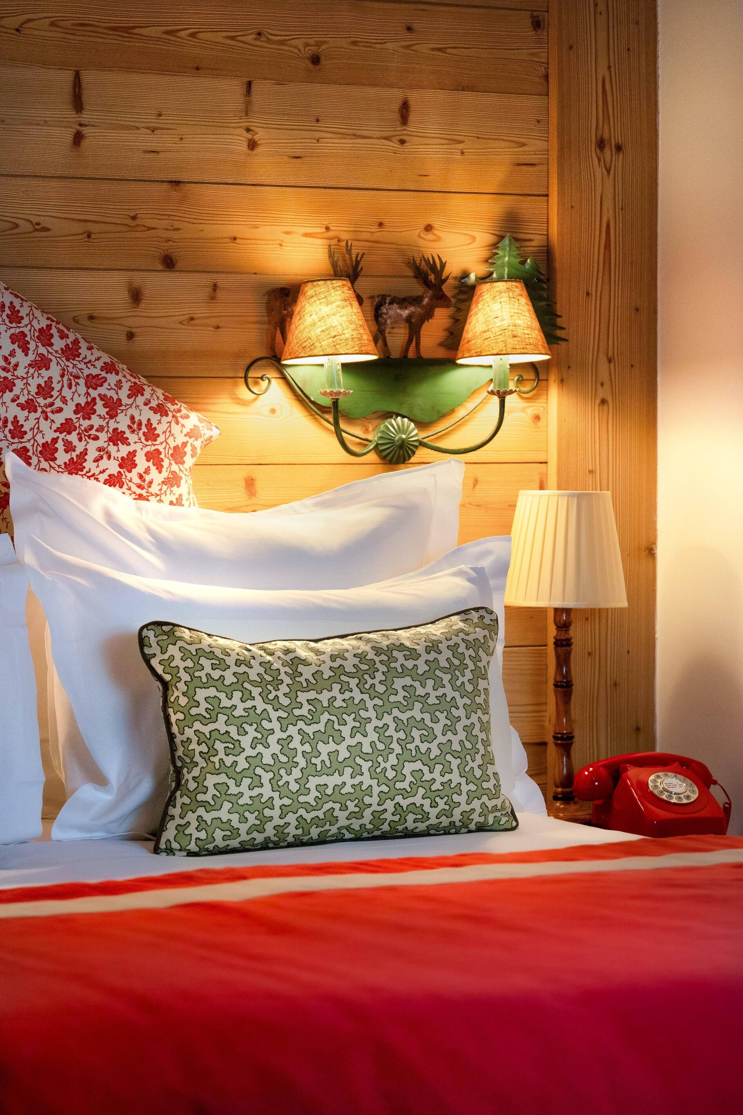 Una cama con una colcha roja, almohadas blancas y almohadas estampadas. Un teléfono rojo está sobre la mesita de noche.