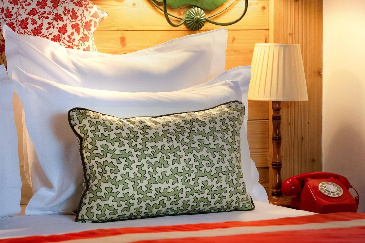 A bed with a red blanket, white pillows, and patterned pillows. A red telephone is on the nightstand.