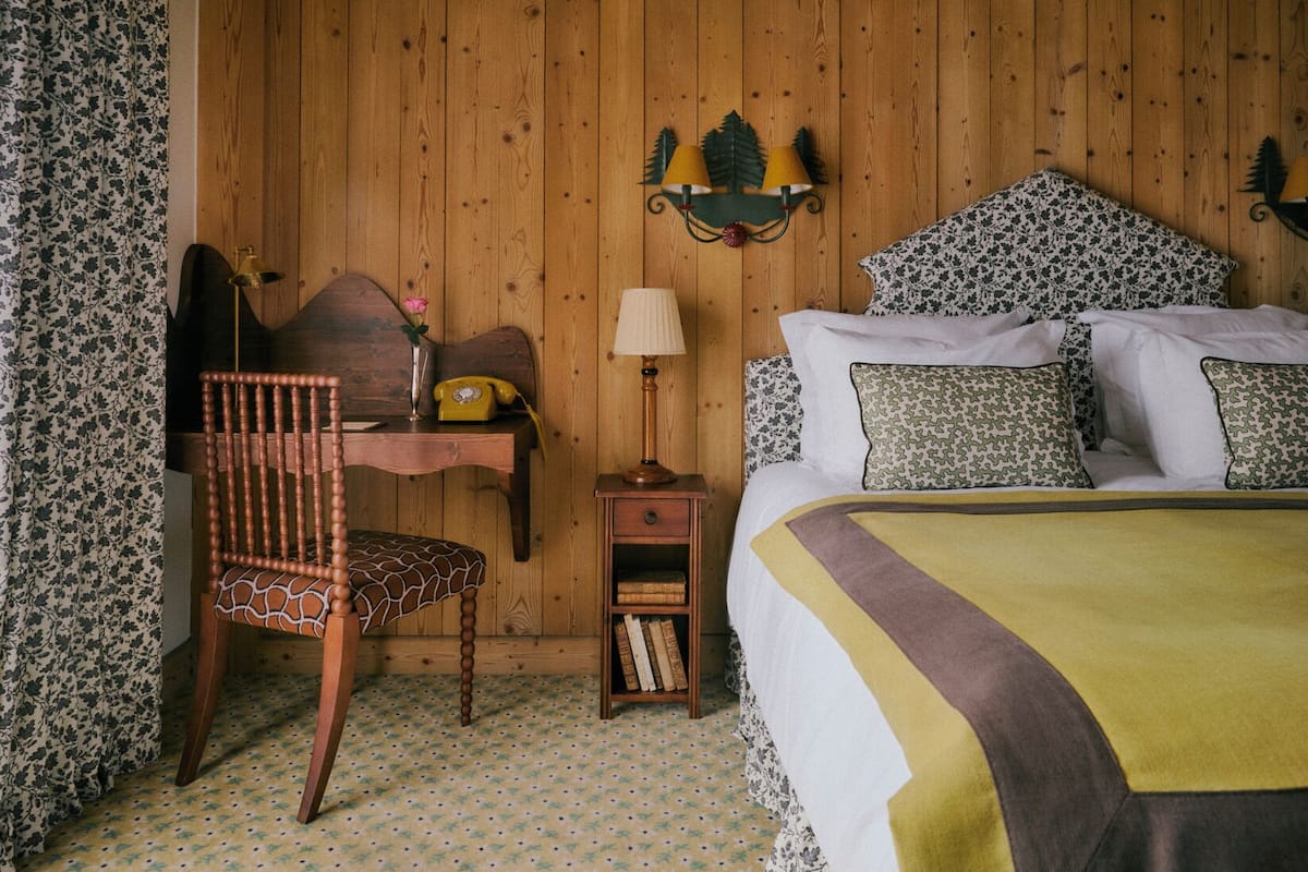 A wooden-paneled room with a bed, a desk with a telephone, a chair, and a lamp.