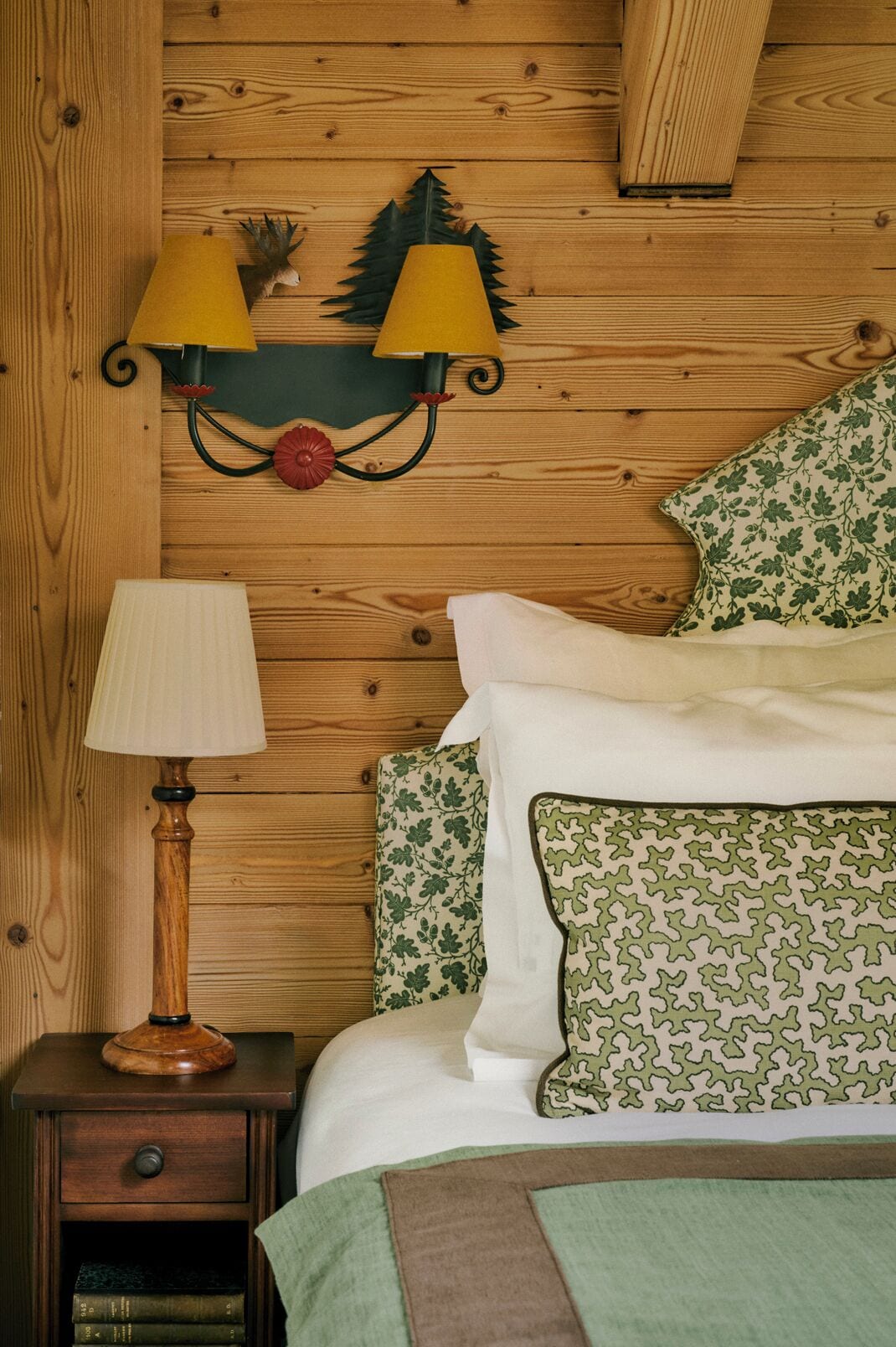 A wooden bedroom with a nightstand, a lamp, a bed with patterned pillows, and a wall-mounted lamp fixture.