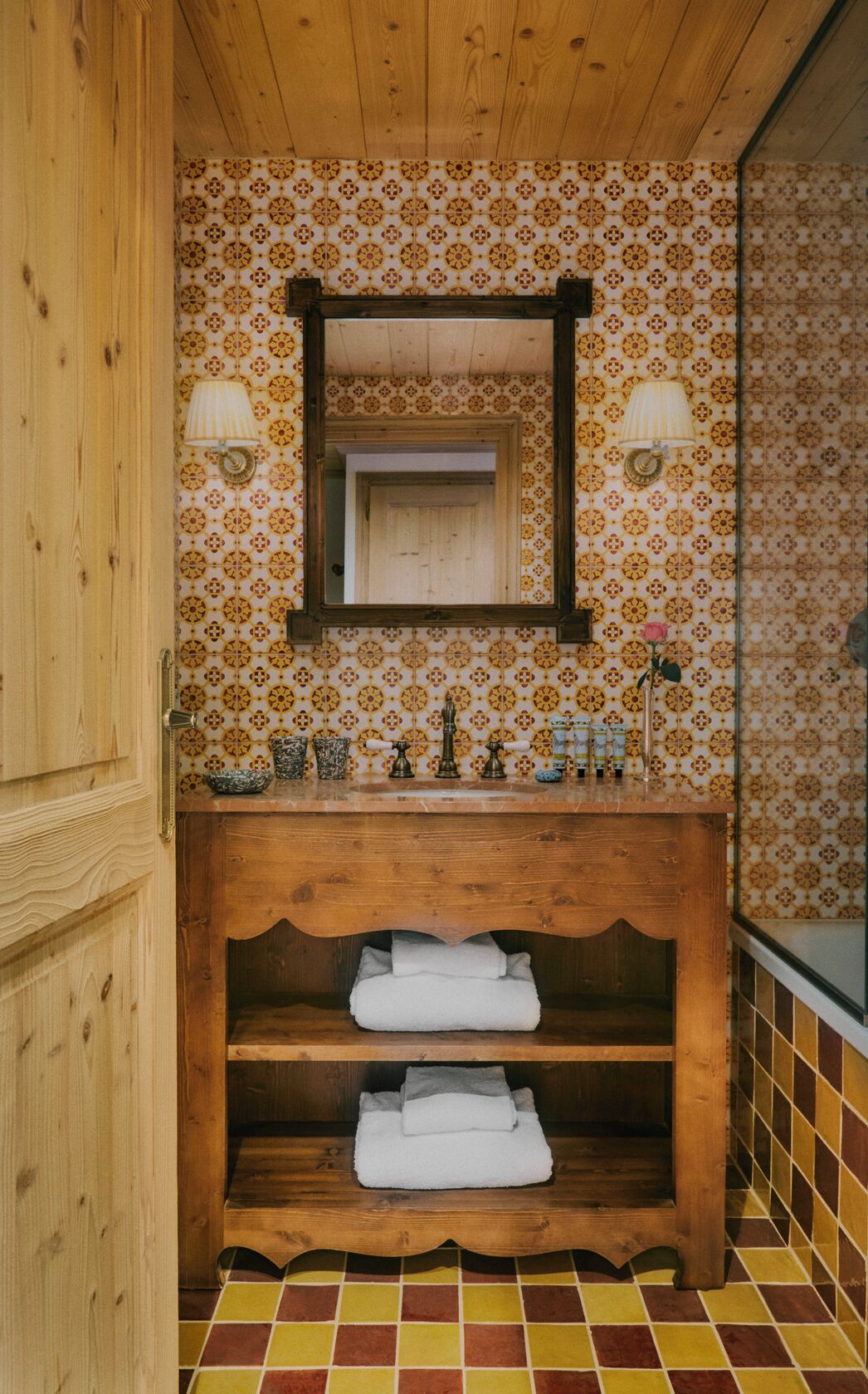 A bathroom with a wooden vanity, a mirror, two wall-mounted lamps, and a tiled floor.