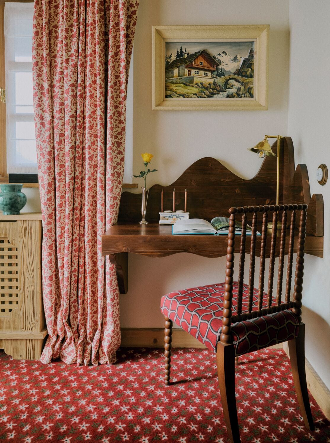 A cozy room with a wooden desk, a chair with a patterned cushion, a framed painting on the wall, and a window with floral curtains.