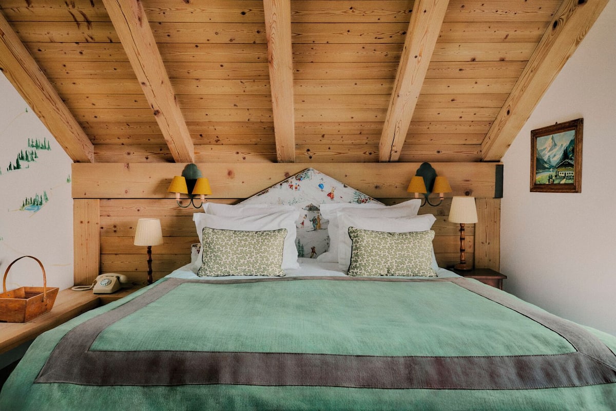A wooden ceiling with exposed beams, a bed with green bedding, two wall-mounted lamps, and a painting on the wall.