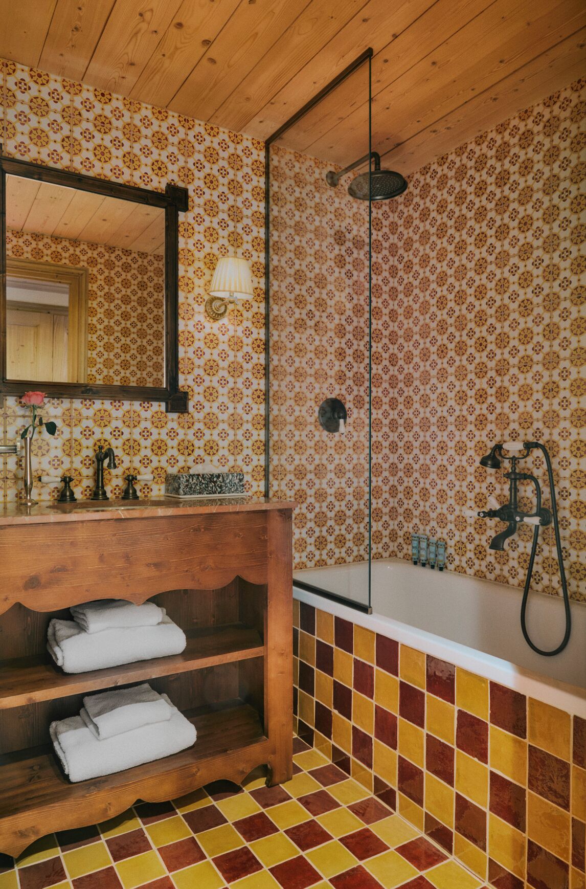 Ein Badezimmer mit einem Holzkosmetikschrank, einem großen Spiegel, einer Dusche inklusive Duschbrausehandstück und einem Fliesenboden.