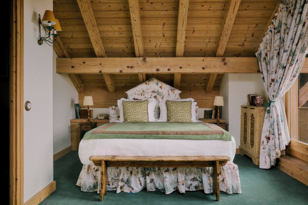 A cozy bedroom with a wooden ceiling, a bed with patterned bedding, a bench, and a nightstand with a lamp.