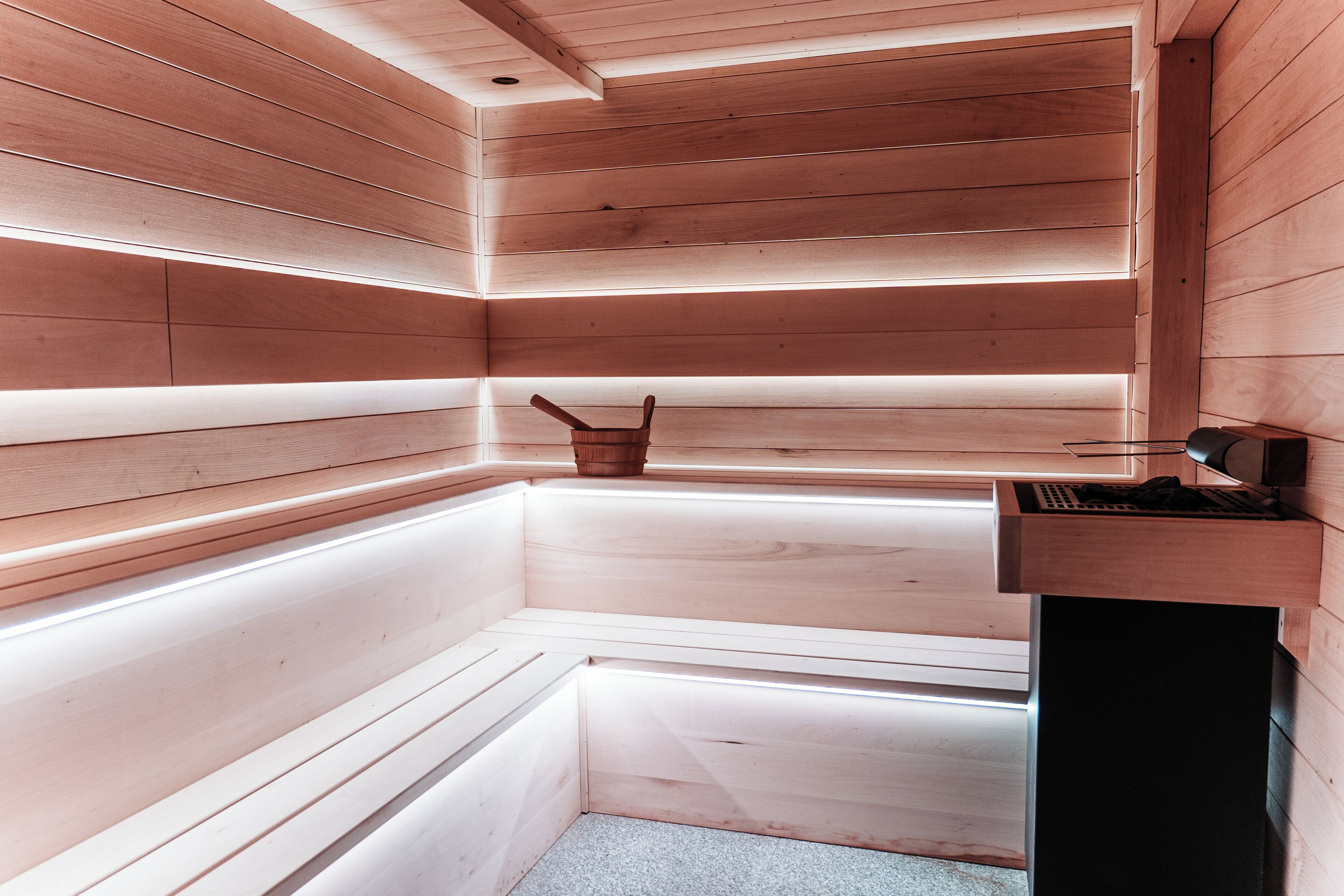 Una sauna con paredes y bancos de madera, un balde pequeño de madera y un calentador negro a la derecha.