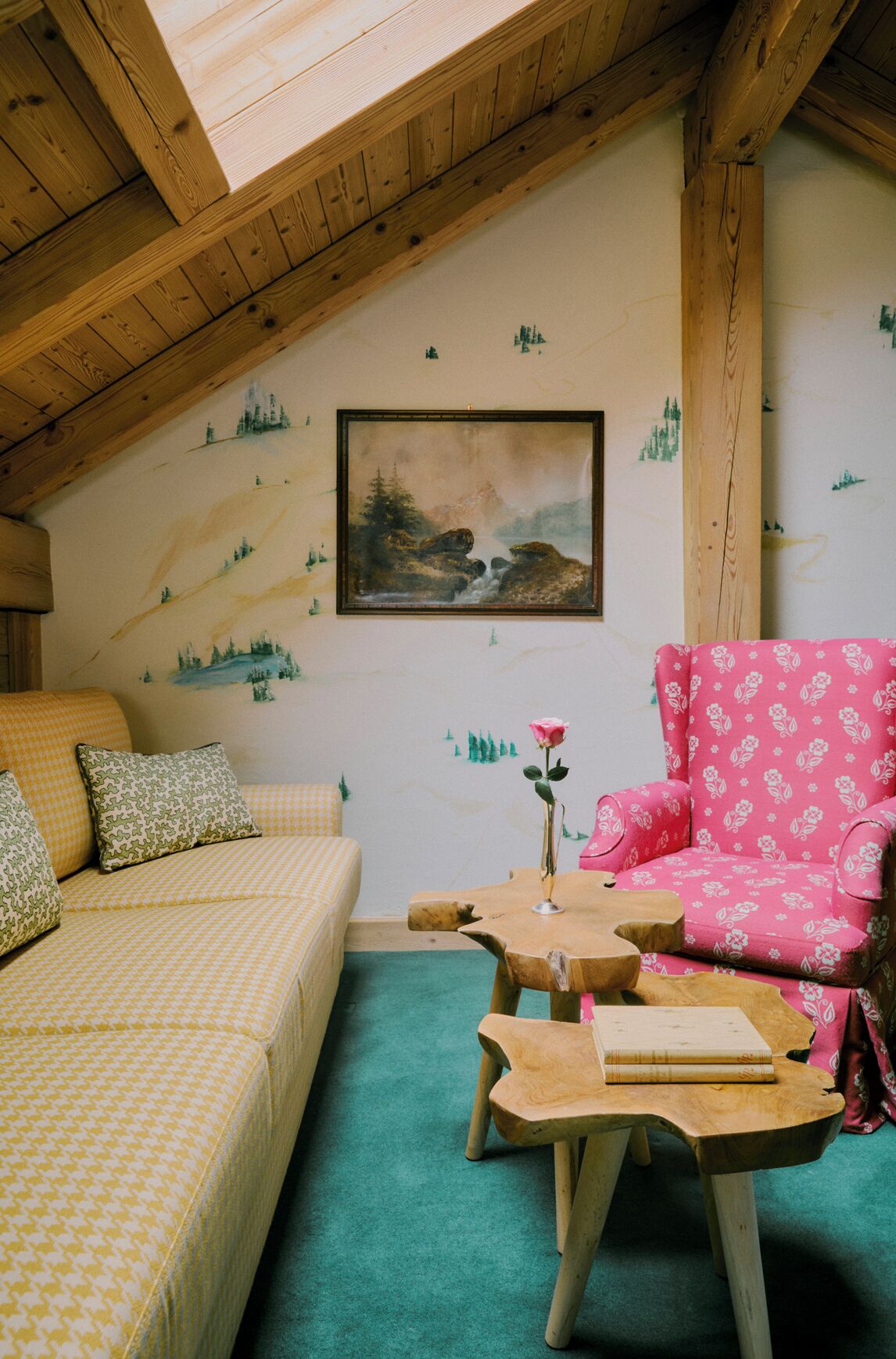 Un cuarto acogedor con techo inclinado, un cuadro de un paisaje montañoso, un sillón floral rosa, un sofá amarillo con estampado y una mesa de centro de madera con un libro.