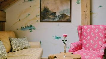 A cozy room with a sloped ceiling, a painting of a mountainous landscape, a pink floral armchair, a yellow patterned sofa, and a wooden coffee table with a book.