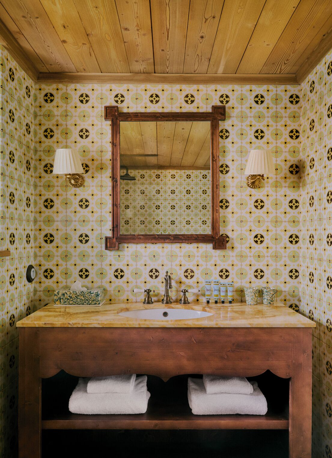 Ein Badezimmer mit einem Holztisch, einem Spiegel, zwei Wandlampen und Wandtapete mit Muster.