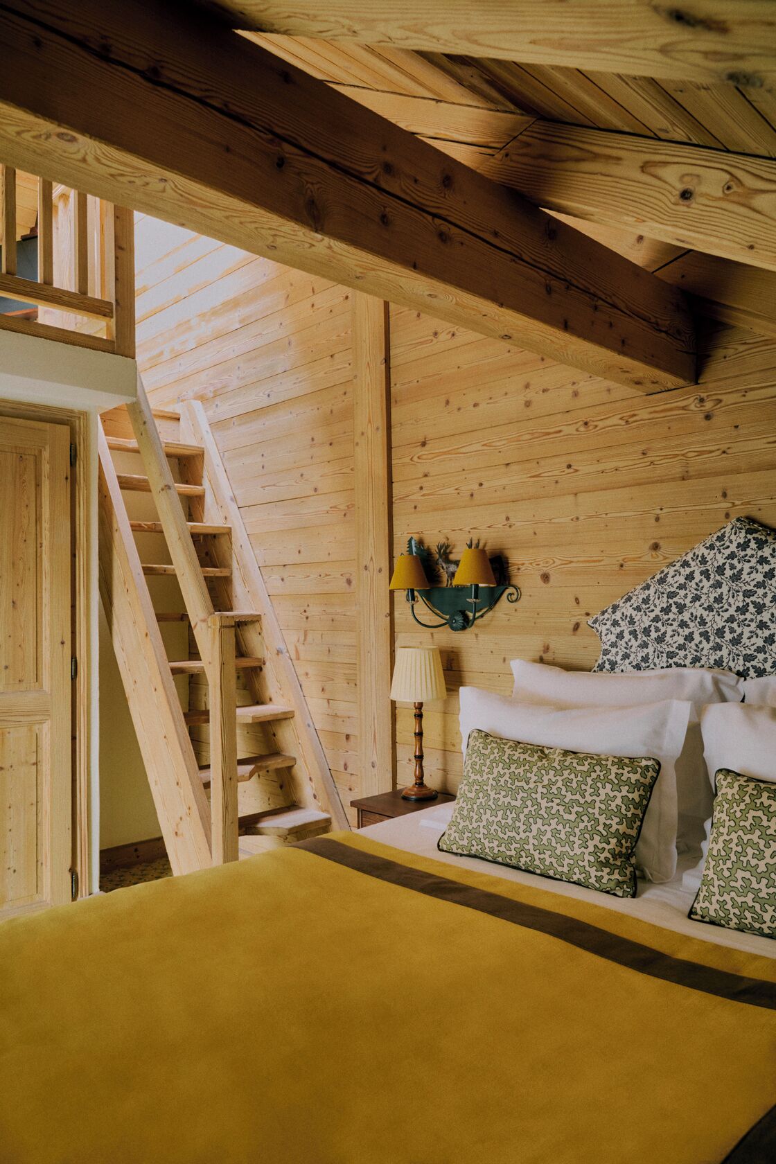 A wooden cabin interior with a bed, a nightstand, a lamp, and a wall-mounted light fixture.