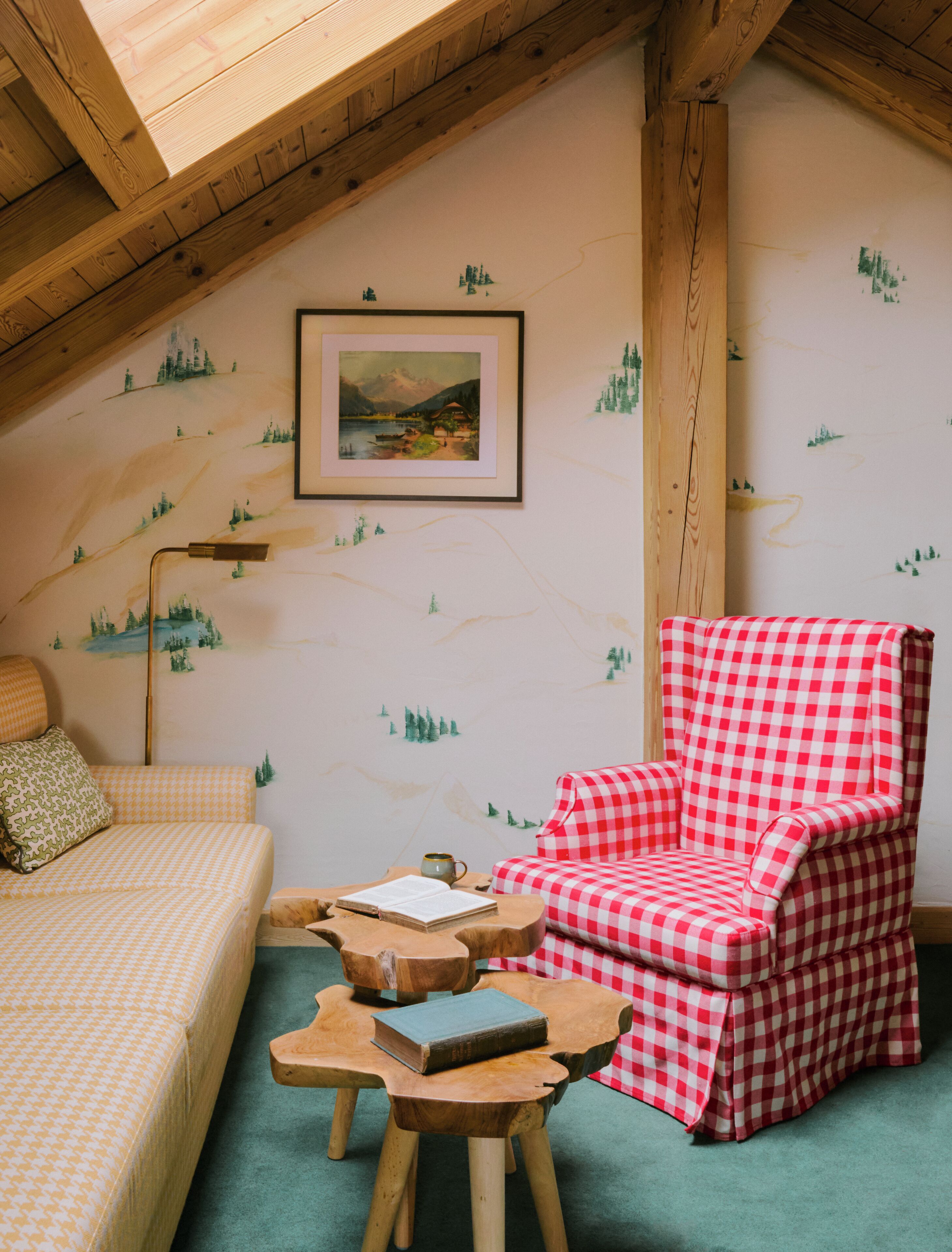 A cozy room with a checkered armchair, a wooden table with books, a bed with a patterned bedspread, and a framed painting on the wall.