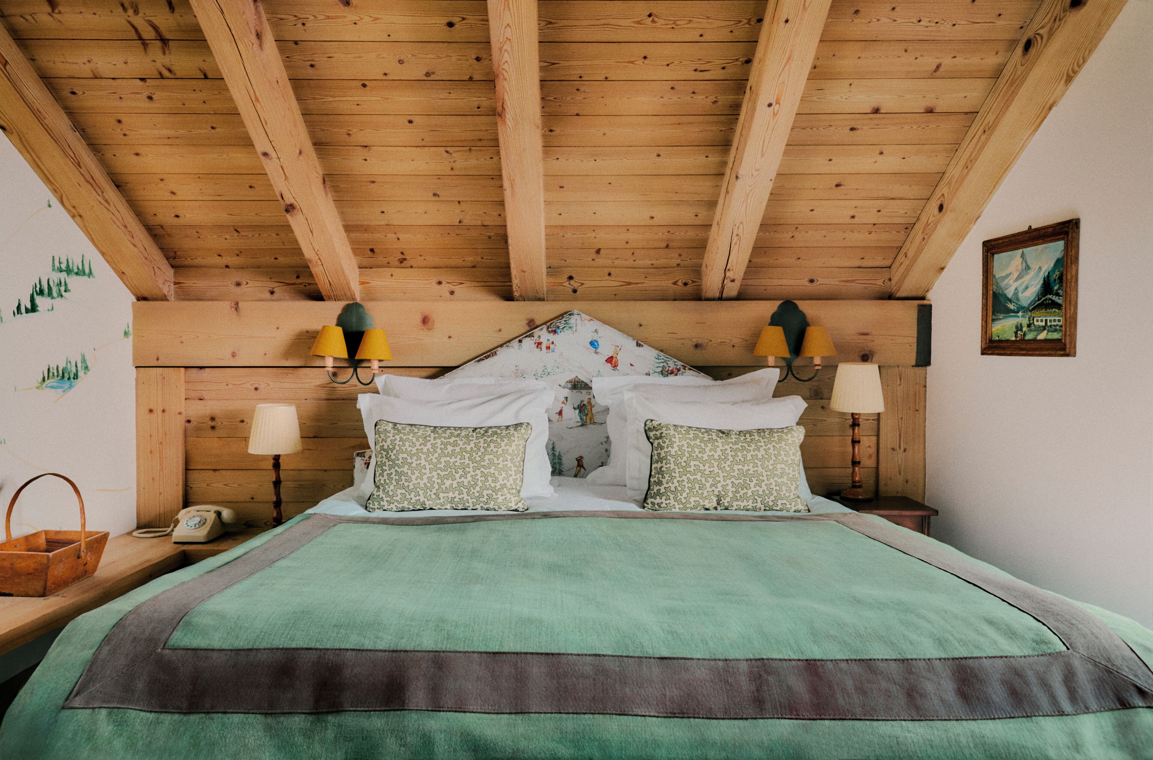 A wooden ceiling with exposed beams, a bed with green bedding, two bedside lamps, a nightstand with a lamp, a telephone, and a wall with a landscape painting.