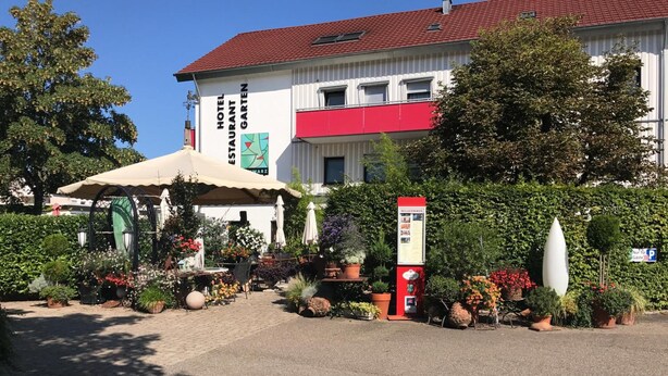 A hotel with a red and white facade, outdoor seating area with umbrellas, and a variety of potted plants. - Hotel Schwarzwälder Hof (Achern)