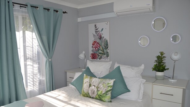 A neatly made bed with a floral pillow, a bedside lamp, and a small potted plant on a white dresser. - Africa Emerald at 47 Petronella (Kempton Park)