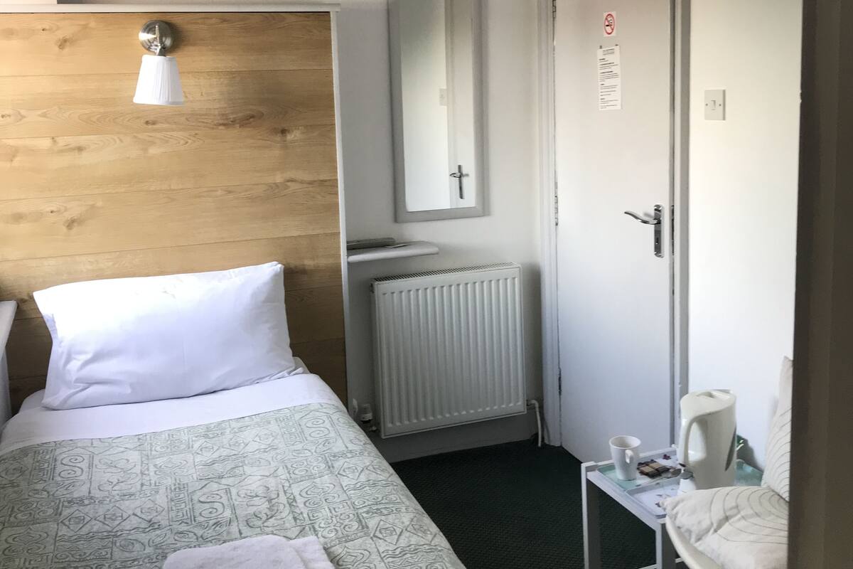 A hotel room with a bed, a wooden headboard, a bedside lamp, a radiator, a small table with a cup and a tissue box, and a door with a sign.