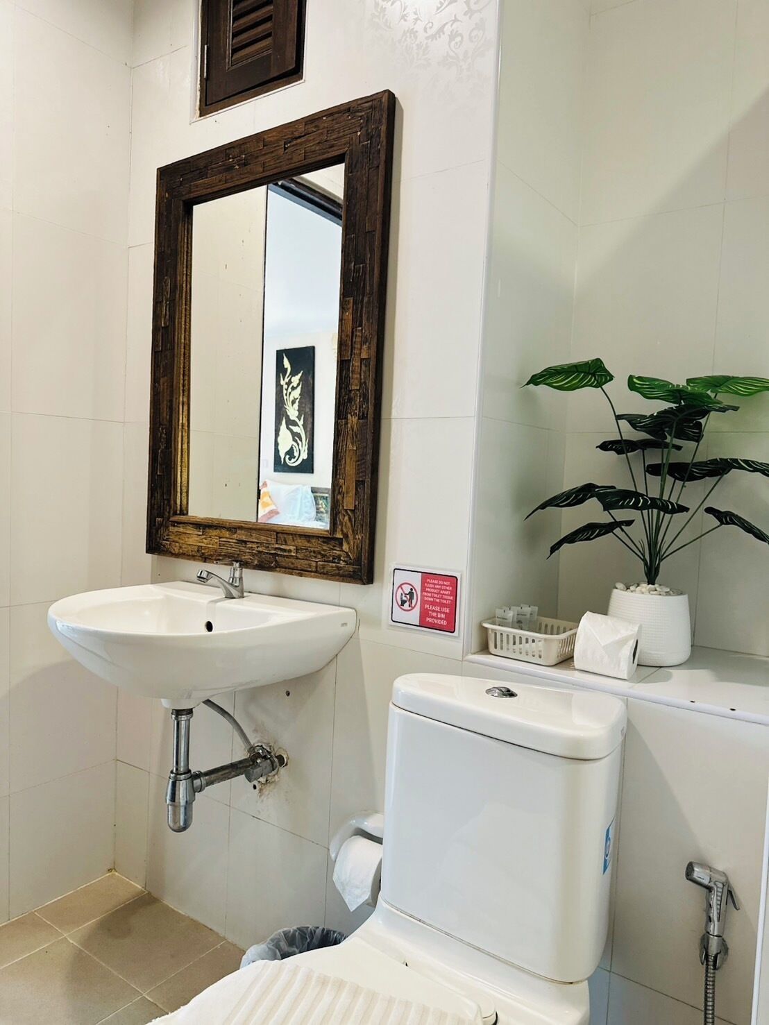A bathroom with a white sink, a mirror with a dark wooden frame, a toilet, a shower, and a potted plant.