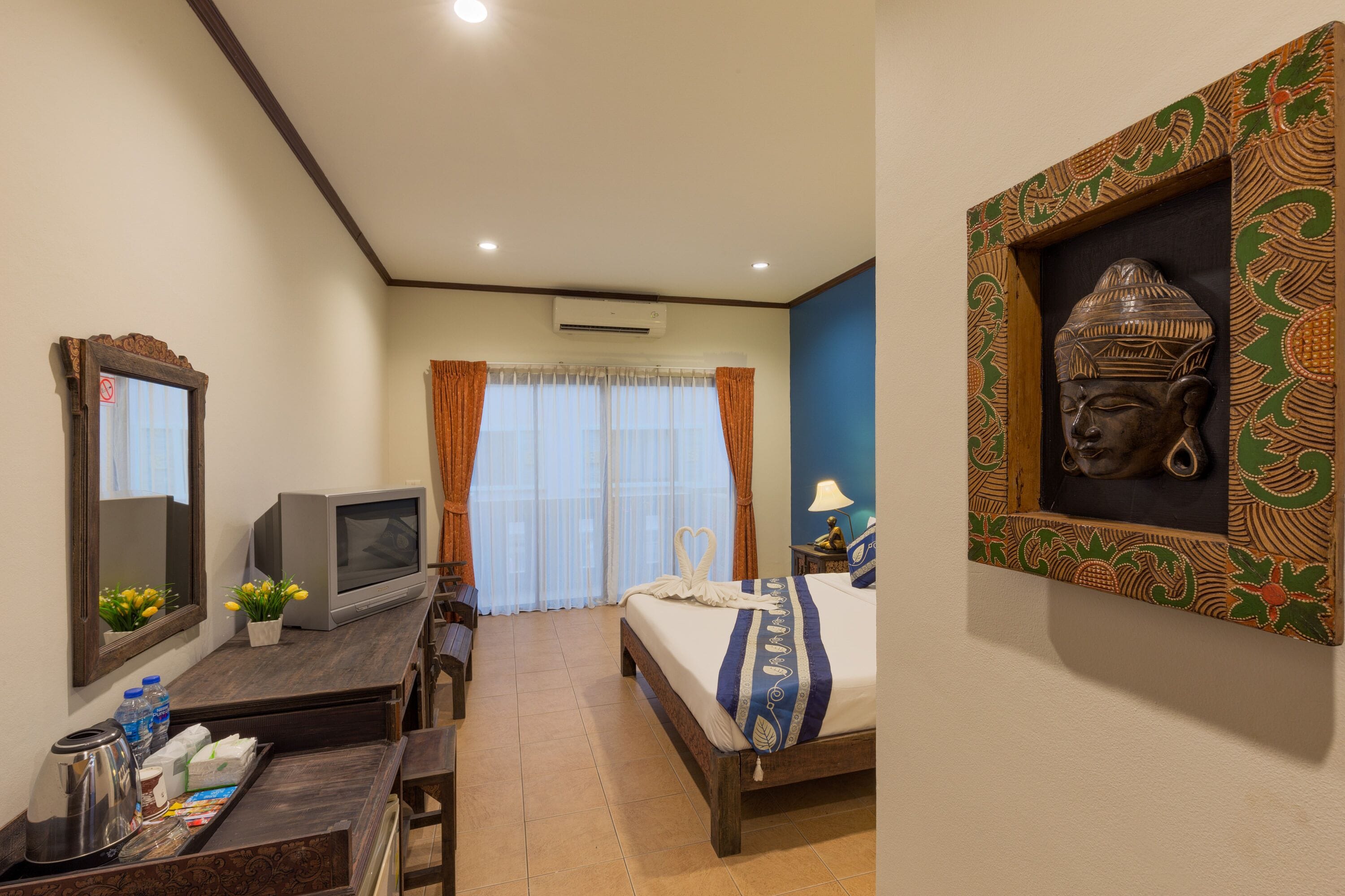 A hotel room with a bed, a desk with a television, a mirror, and a Buddha statue on the wall.