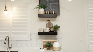 Uma cozinha com armários brancos, uma prateleira preta com objetos decorativos, um lustre e uma frase inspiradora emoldurada na parede.