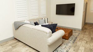 Uma sala de estar com um sofá bege, um banquinho marrom e uma grande televisão de tela plana na parede.