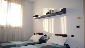 A modern bedroom with two beds, a shelf above, and a window with curtains. - BNBOOK-RESIDENZA NAGGI (CASORATE SEMPIONE)