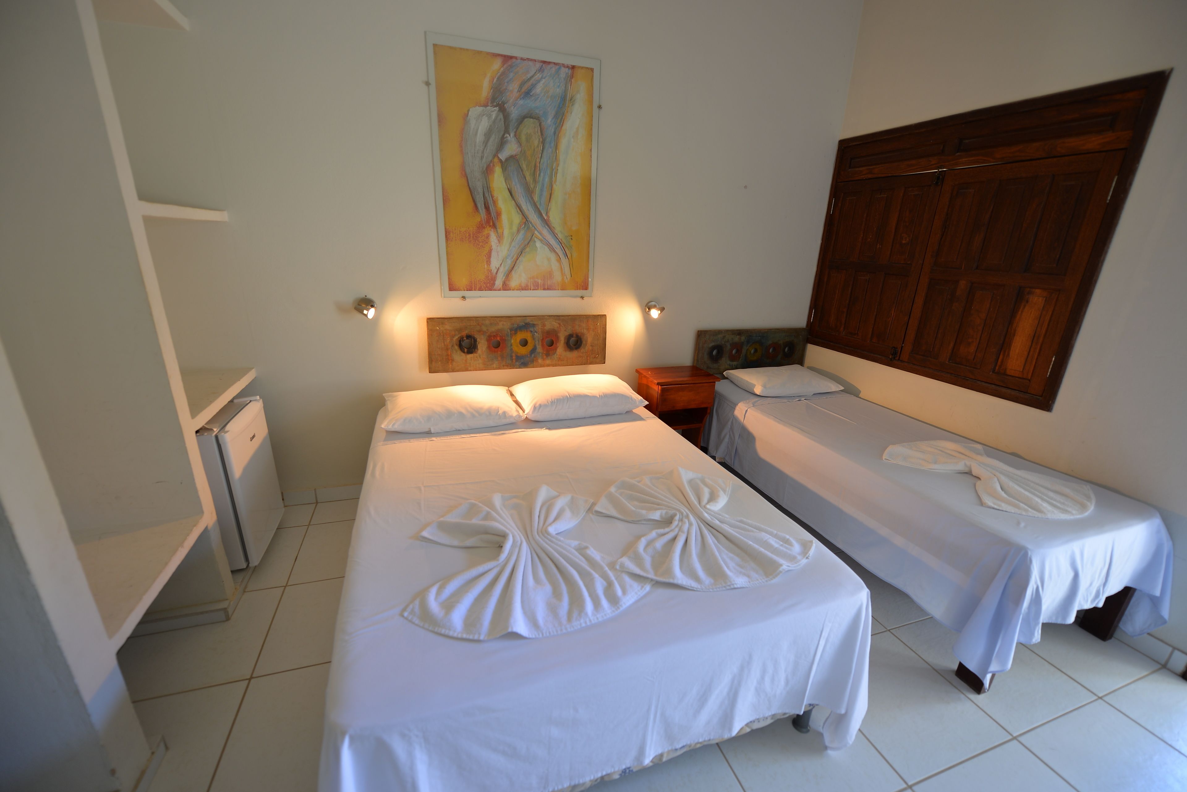 A hotel room with two beds, a wooden wardrobe, a painting on the wall, and a small desk with a lamp.