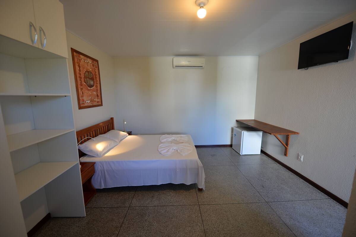A bedroom with a bed, a bedside table, a shelf unit, an air conditioning unit, and a wall-mounted TV.