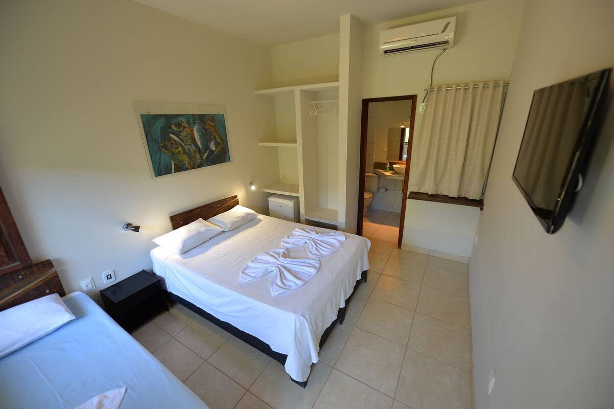 A hotel room with a large bed, a bedside table, a nightstand, a painting on the wall, an air conditioning unit, and a bathroom visible through an open door.
