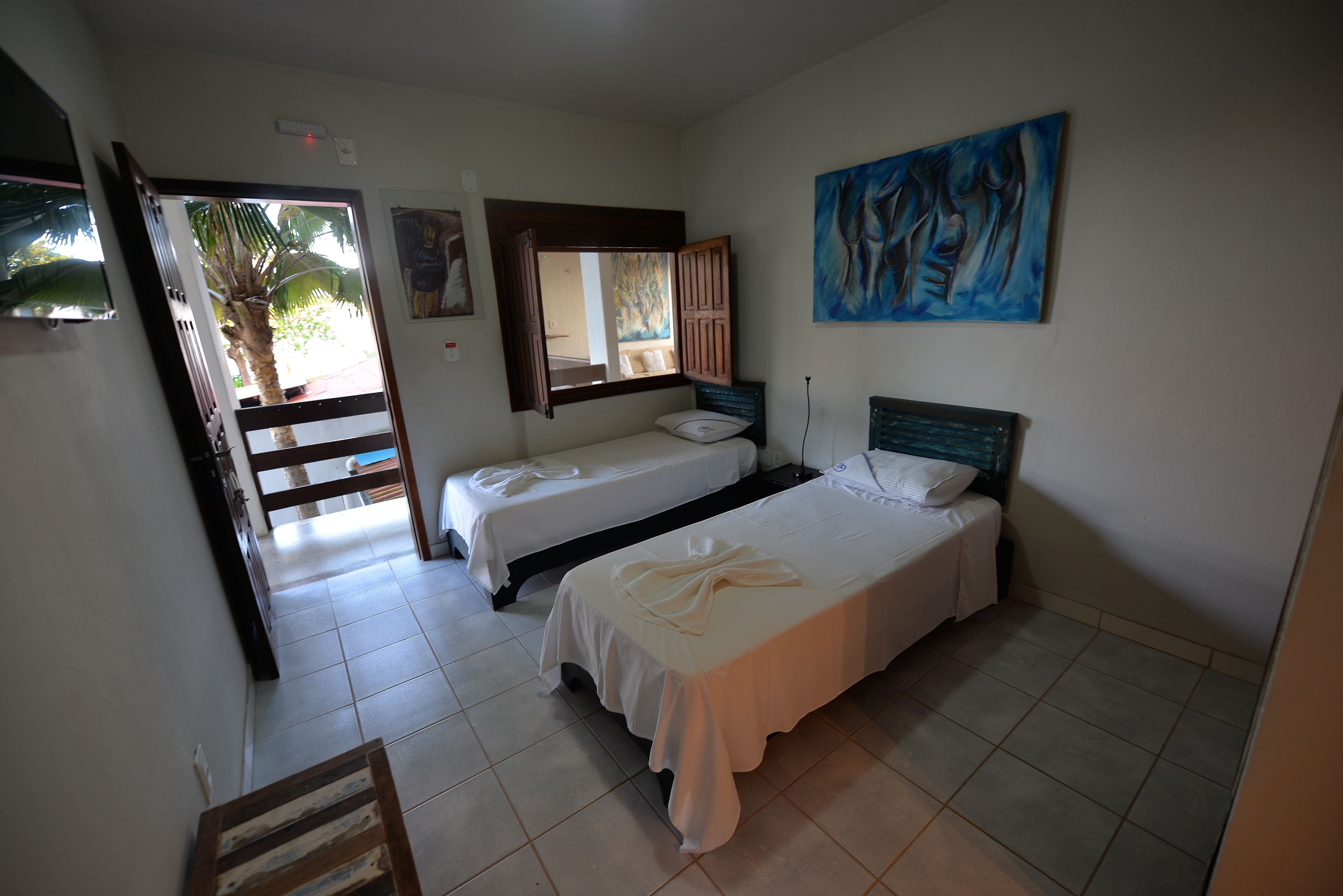 A room with two single beds, a large mirror, a painting, and a view of a palm tree through an open door.