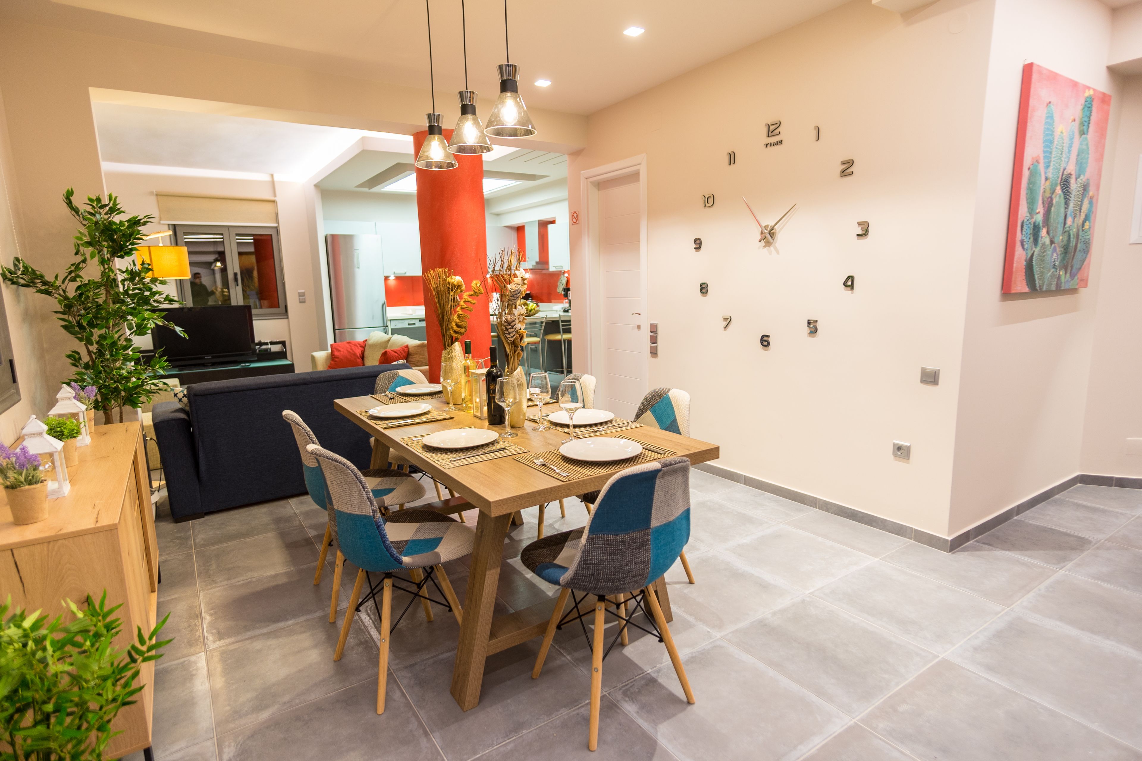 Een eethoek met een houten tafel gedekt voor vier personen, omringd door blauwe en grijze stoelen. Aan de muur hangt een grote klok en rechts een schilderij van een cactus.
