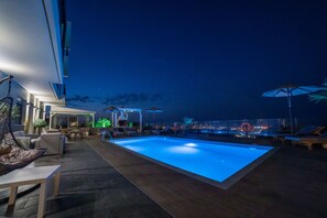 A nighttime swimming pool area with lounge chairs and umbrellas, a covered seating area with tables, and a view of the city lights in the background. - Villa Constandinos (Hersonissos)