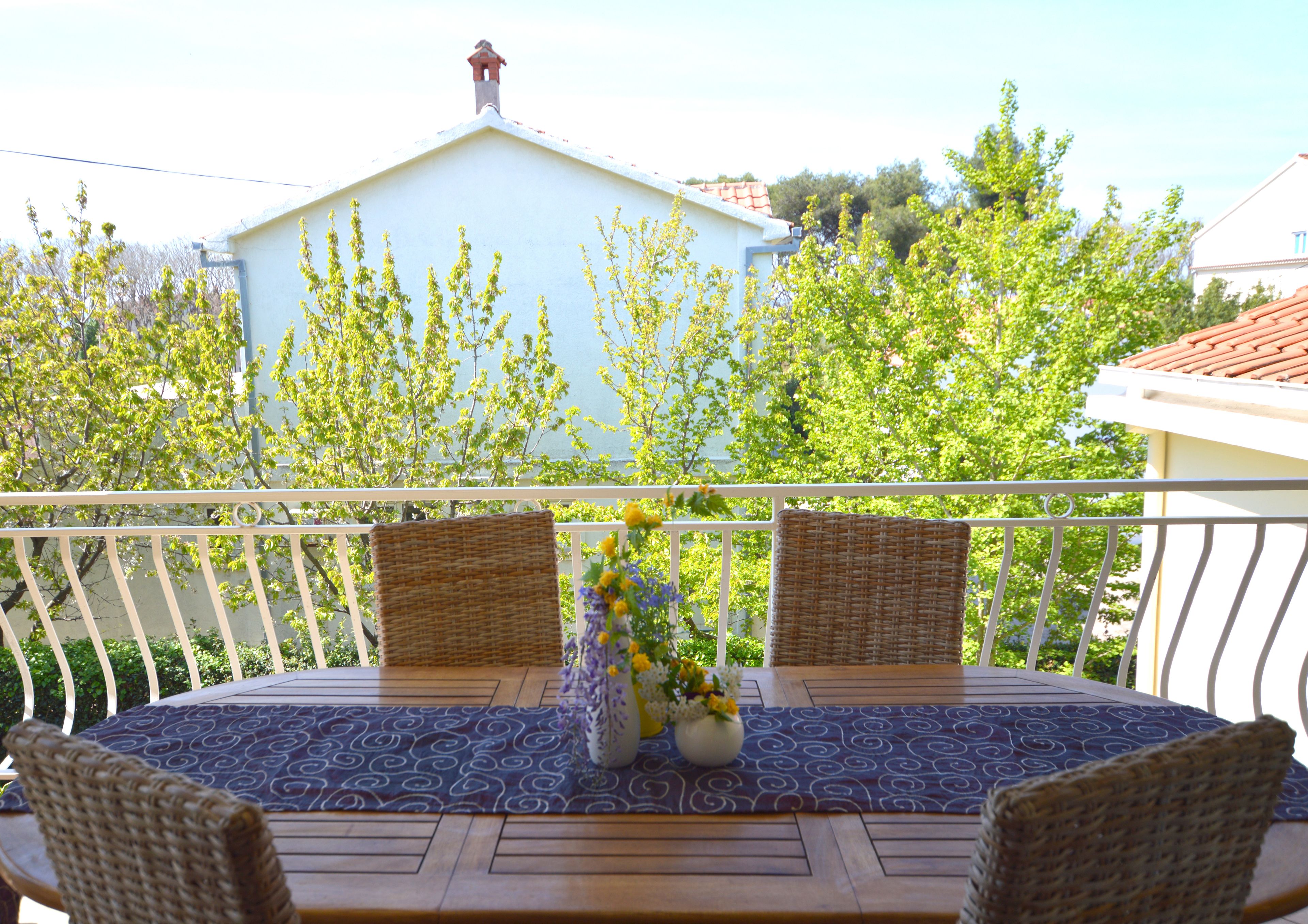 Ein Balkon mit Tisch für zwei Personen, zwei Korbstühlen und einem Blumenvasen.