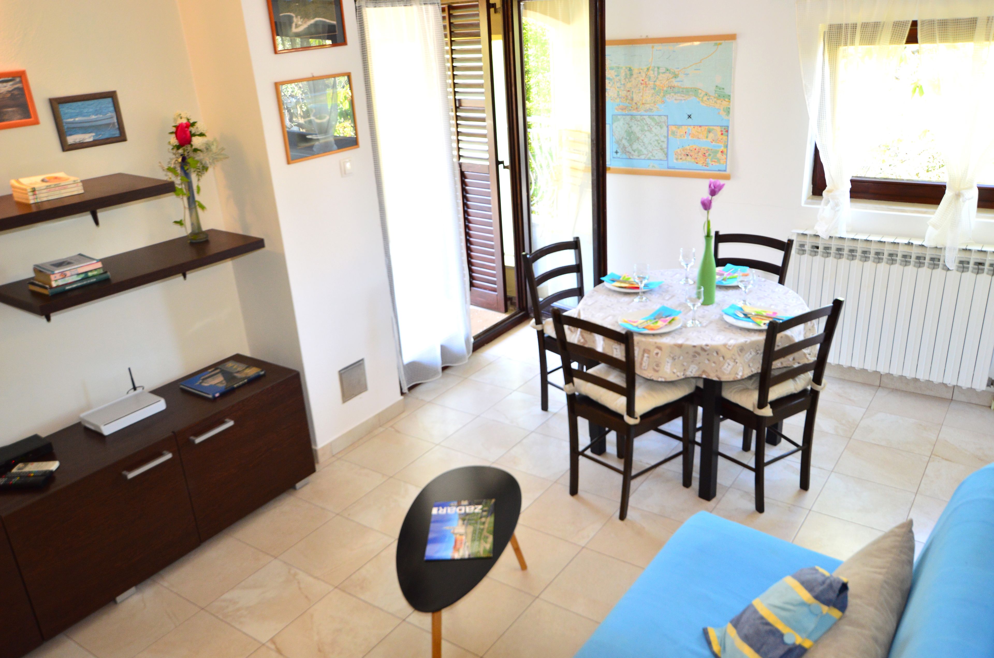 A dining area with a table, chairs, and a map on the wall. There is a sofa and a cabinet with books and a vase of flowers.