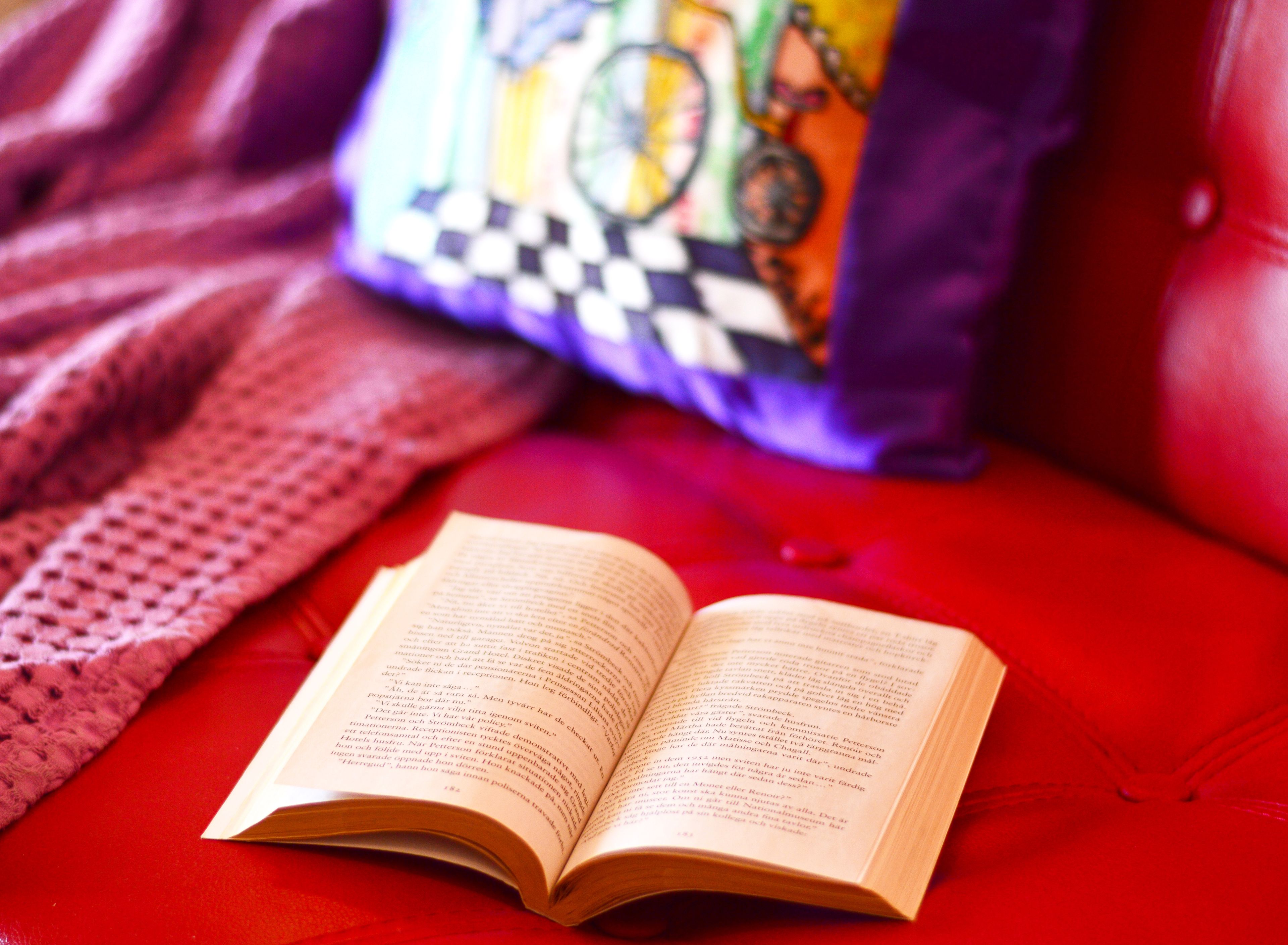 An open book on a red surface with a colorful pillow and a knitted blanket in the background.