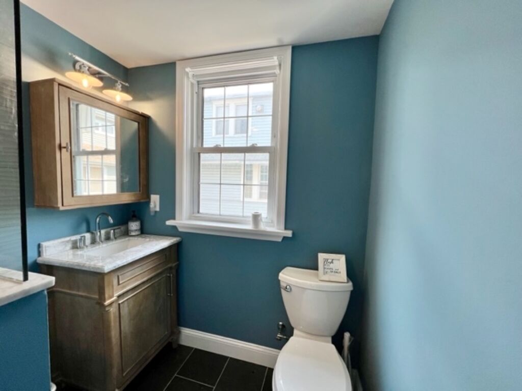 Een badkamer met een blauwe wand, een houten wastafelmeubel, een wit toilet en een raam boven het toilet.