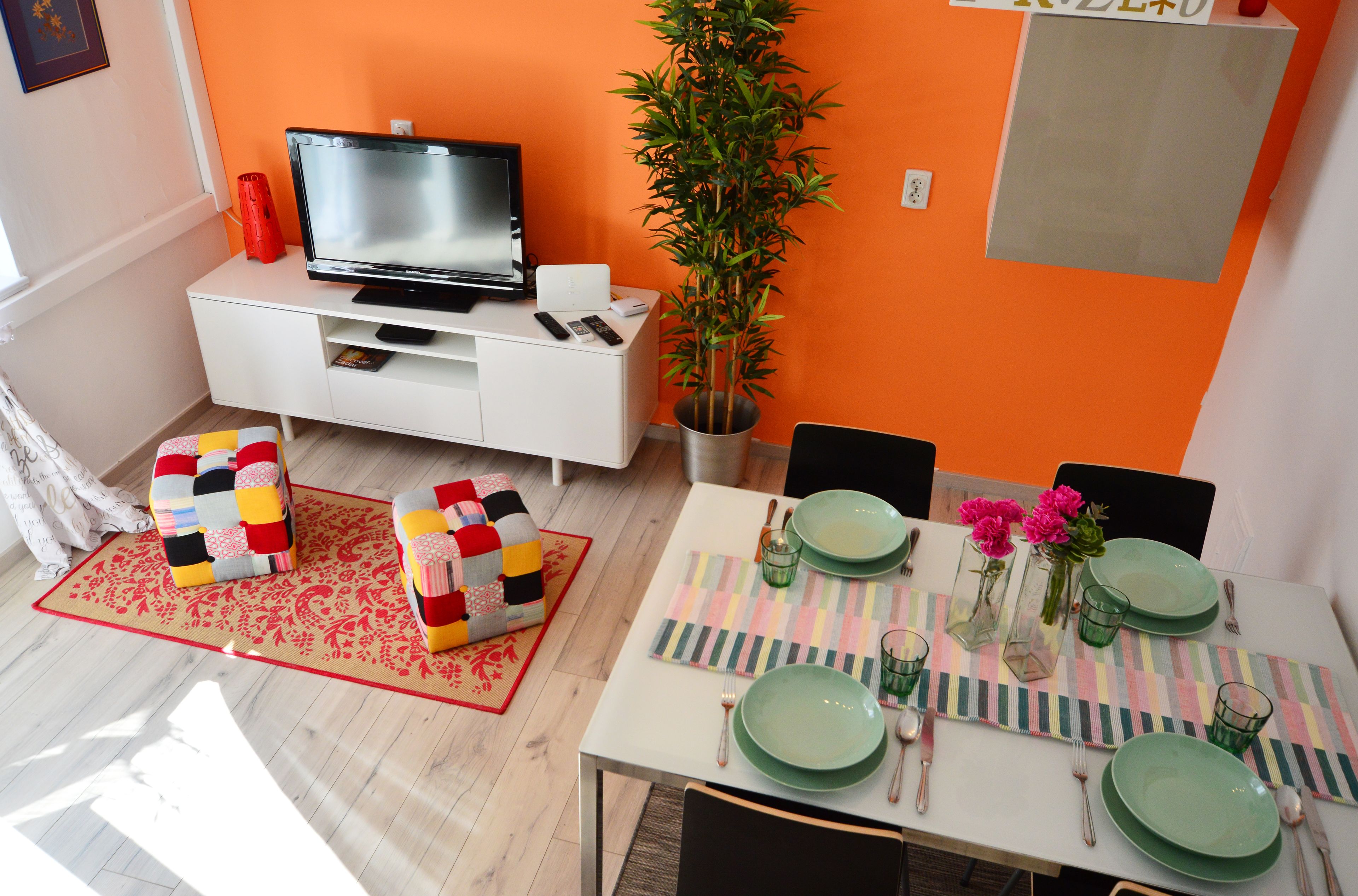 A dining area with a table set for four, a television on a white cabinet, and a potted plant.