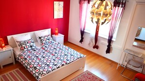 Un dormitorio con cama, mesitas de noche, cómoda, ventana con cortinas y lámpara colgante.