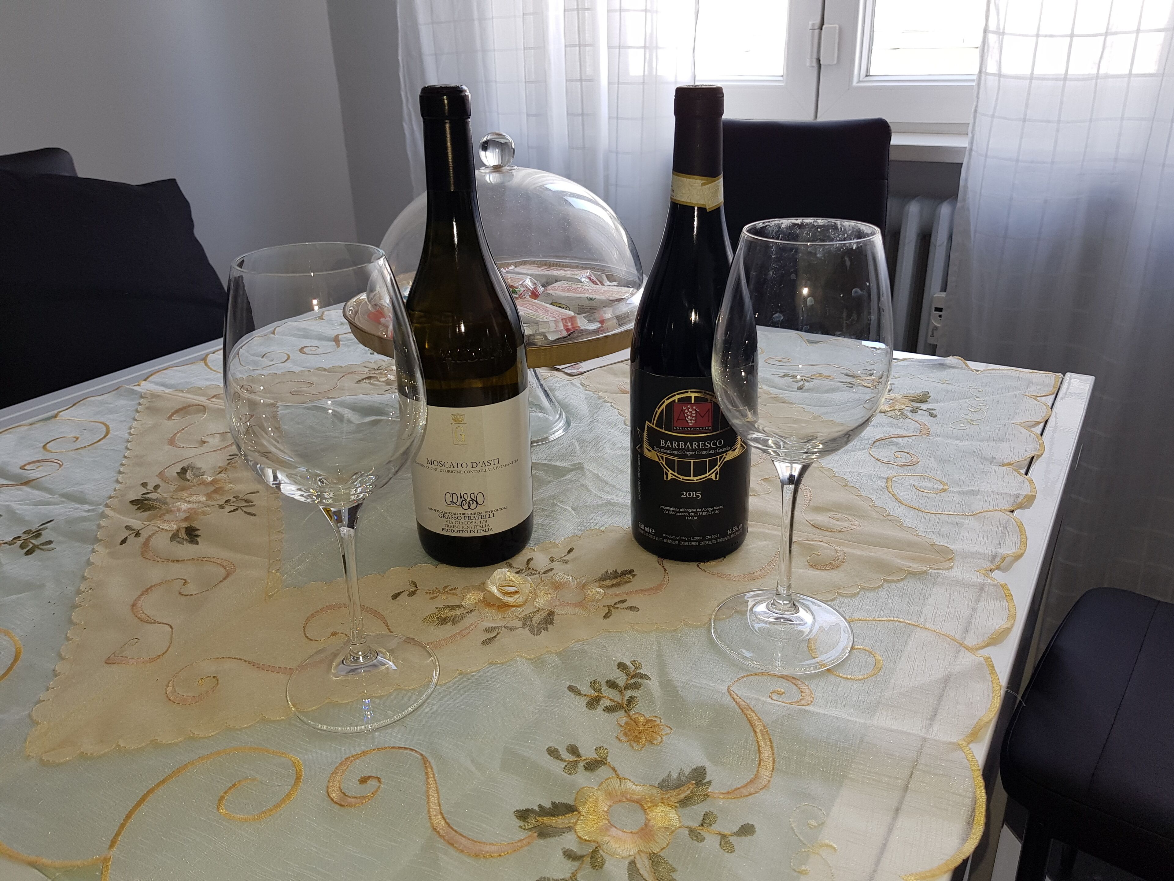 Une table à manger avec deux bouteilles de vin et deux verres, une nappe décorative et une fenêtre avec des rideaux.