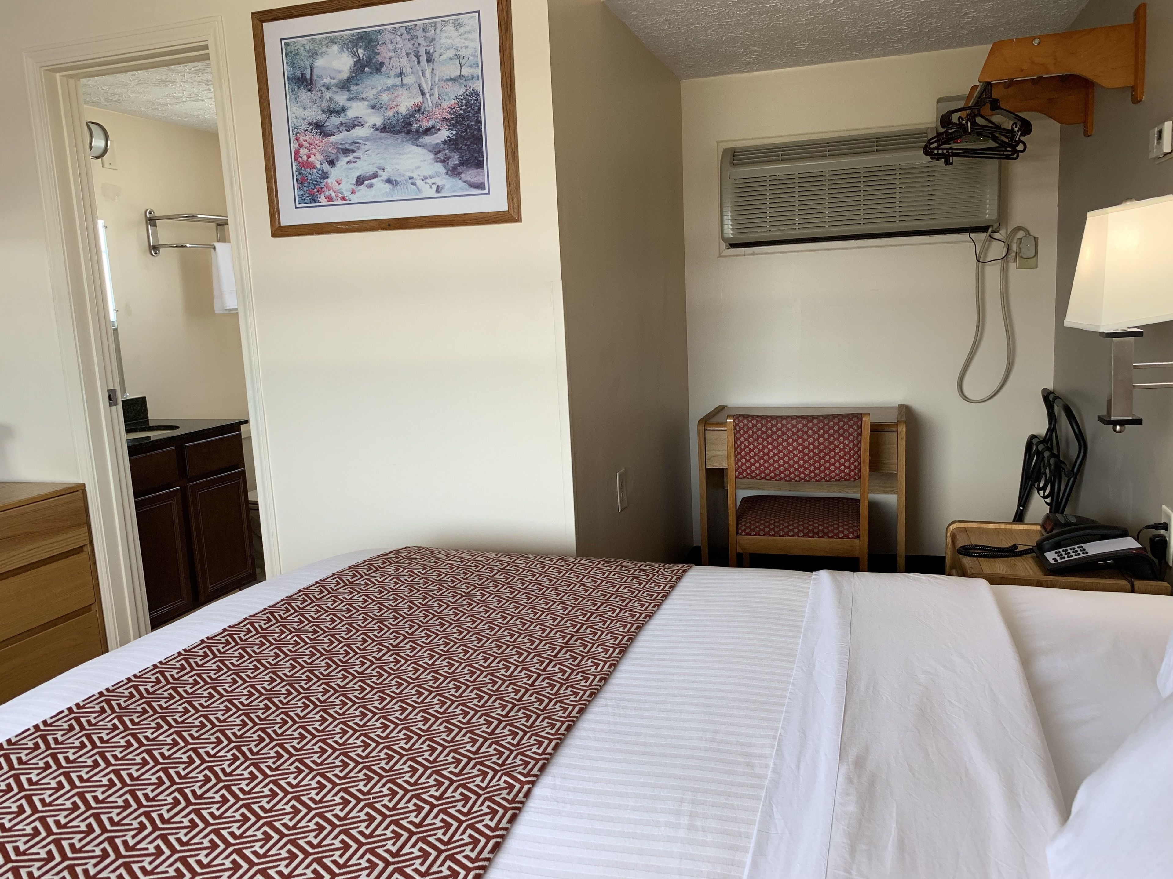 Une chambre d’hôtel avec un lit, une chaise en bois, un bureau avec une lampe et un tableau au mur.