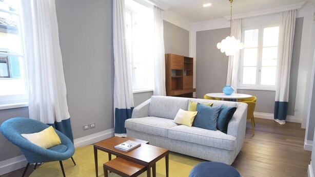 Un salon avec un canapé gris, un fauteuil bleu et un pouf rond. On y trouve une table basse en bois et un tapis jaune.