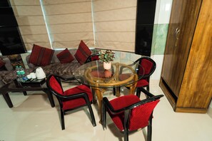 A dining area with a round glass table surrounded by red upholstered chairs, a sofa with patterned cushions, and a wooden cabinet. - Vrinda Palace (Mathura)
